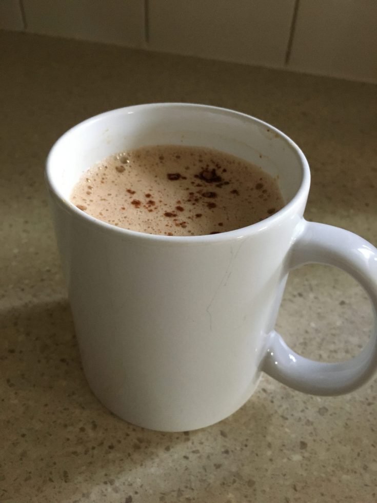Mug with frothy coffee and a sprinkle of cocoa on counter top. Sugar-free Keto peanut butter coffee recipe with Hershey's cocoa.