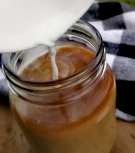 A pumpkin spice latte is being made. Milk is poured from a small white bowl into a clear mason jar filled with coffee. The mason jar is on a wooden surface and a black and white plaid towel is visible in the background.