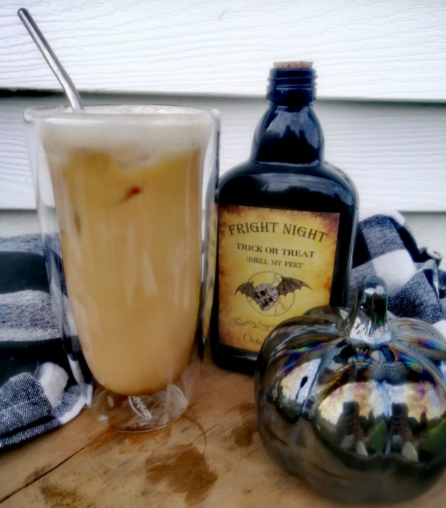 Iced pumpkin spice latte with whipped cream. The iced pumpkin spice latte is in a clear glass with a metal straw. The Latte is on a wooden surface with a black glass bottle that reads "Fright night." That bottle has a black glass pumpkin in front of it. There is a black and white checkered towel in the background along with a white background overall.