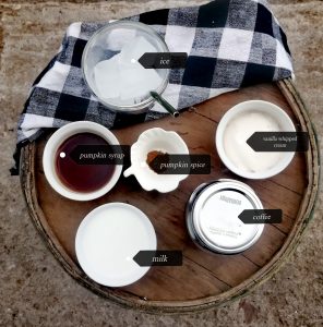 There are 6 small white bowls with the following ingredients inside them "Ice, vanilla whipped cream, coffee, milk, pumpkin syrup, and pumpkin spice" the bowls are on a wooden surface with a black and white checkered towel in the background and the background is grey overall. 