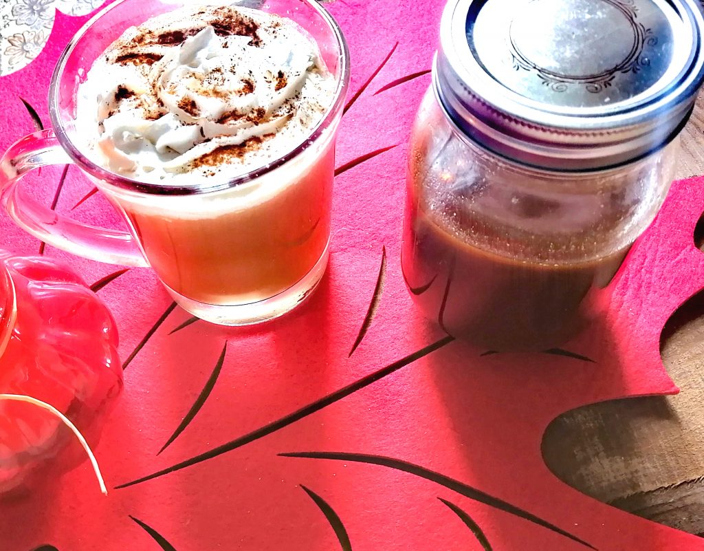 Pumpkin spice latte in a clear glass mug, topped with whipped cream and a sprinkle of pumpkin spice beside a mason jar with a silver lid filled with pumpkin spice syrup. Both objects are on a large red maple leaf with a wooden surface beneath it.
